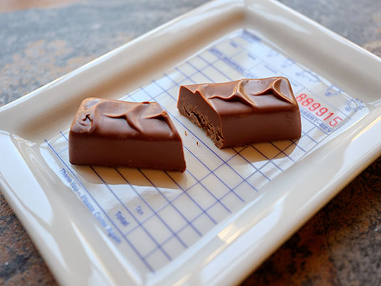 Two pieces of chocolate on a white tray with a grid background Ara Chocolat Sesame Praline Bar