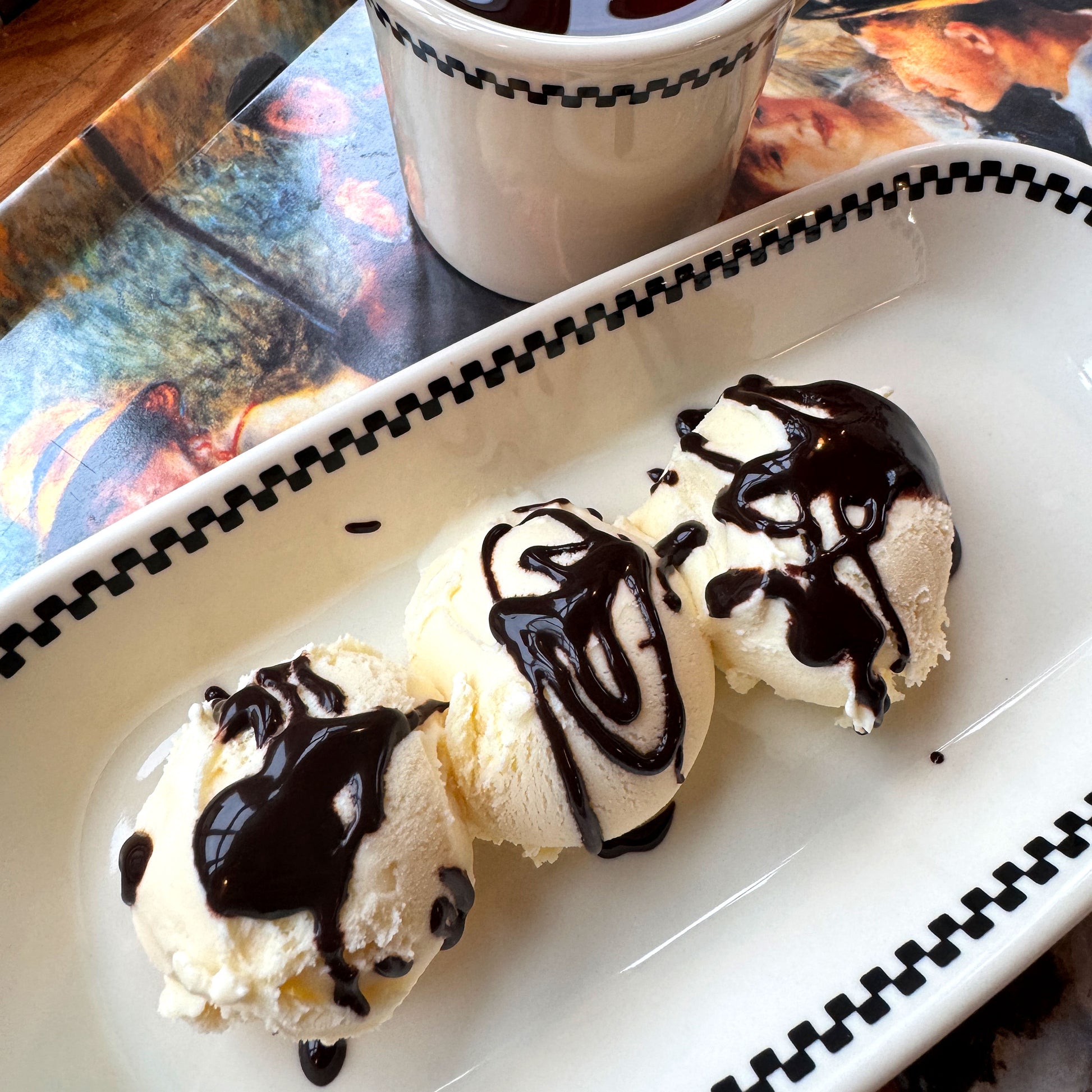 Three scoops of ice cream with chocolate drizzle on a white tray with black checkered border, next to a cup of coffee. ChefShop Grande Brut Cocoa Powder