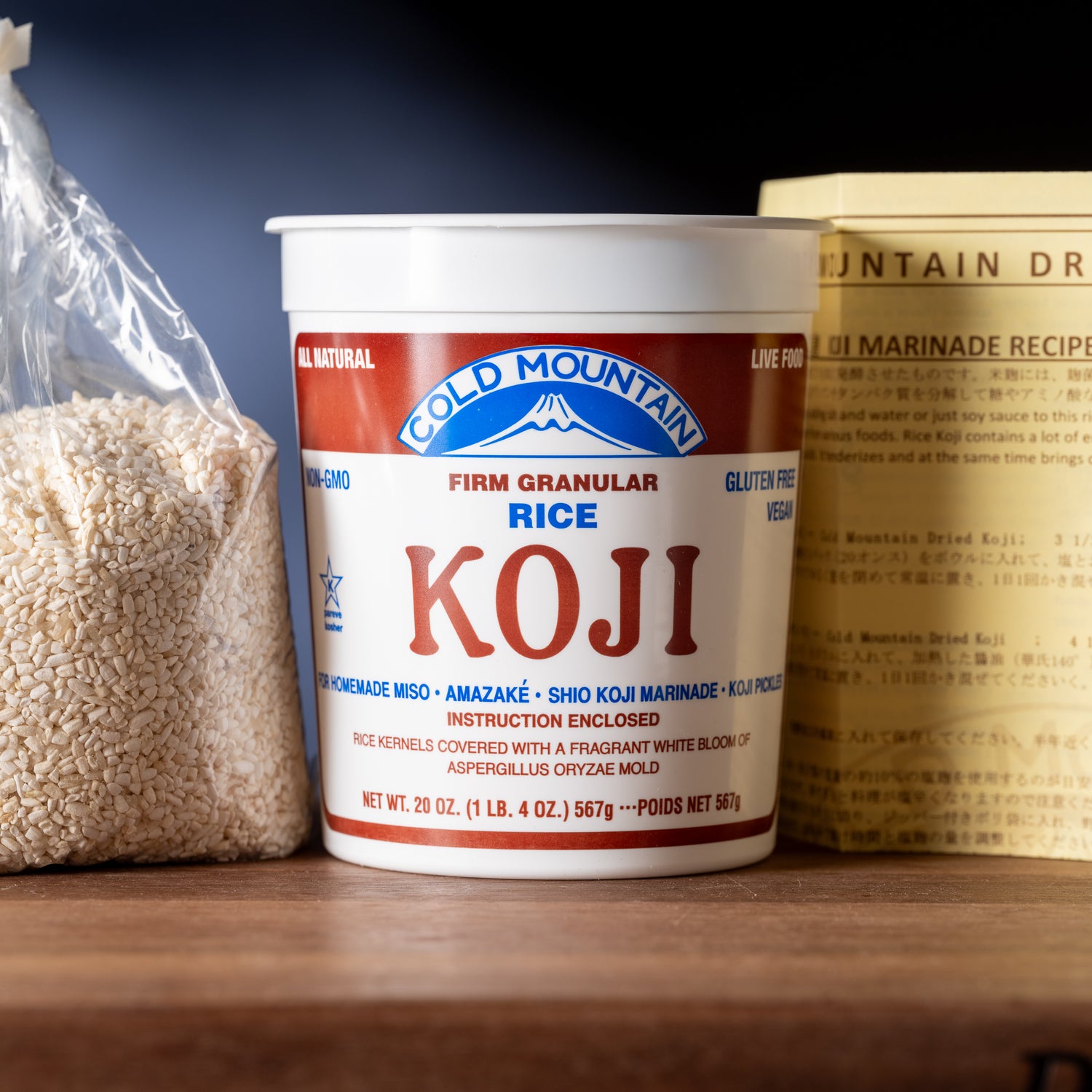 Container of Gold Mountain Koji rice on a wooden surface with rice and another product in the background.
