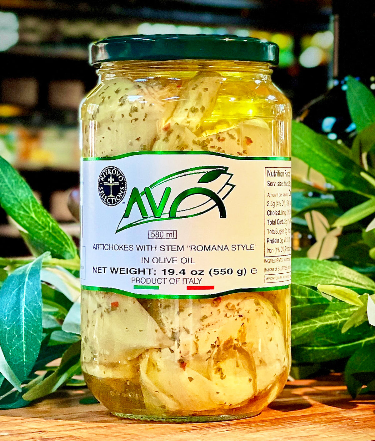 A glass jar of AVA brand artichokes with stem in olive oil, set on a wooden surface with green leaves in the background.