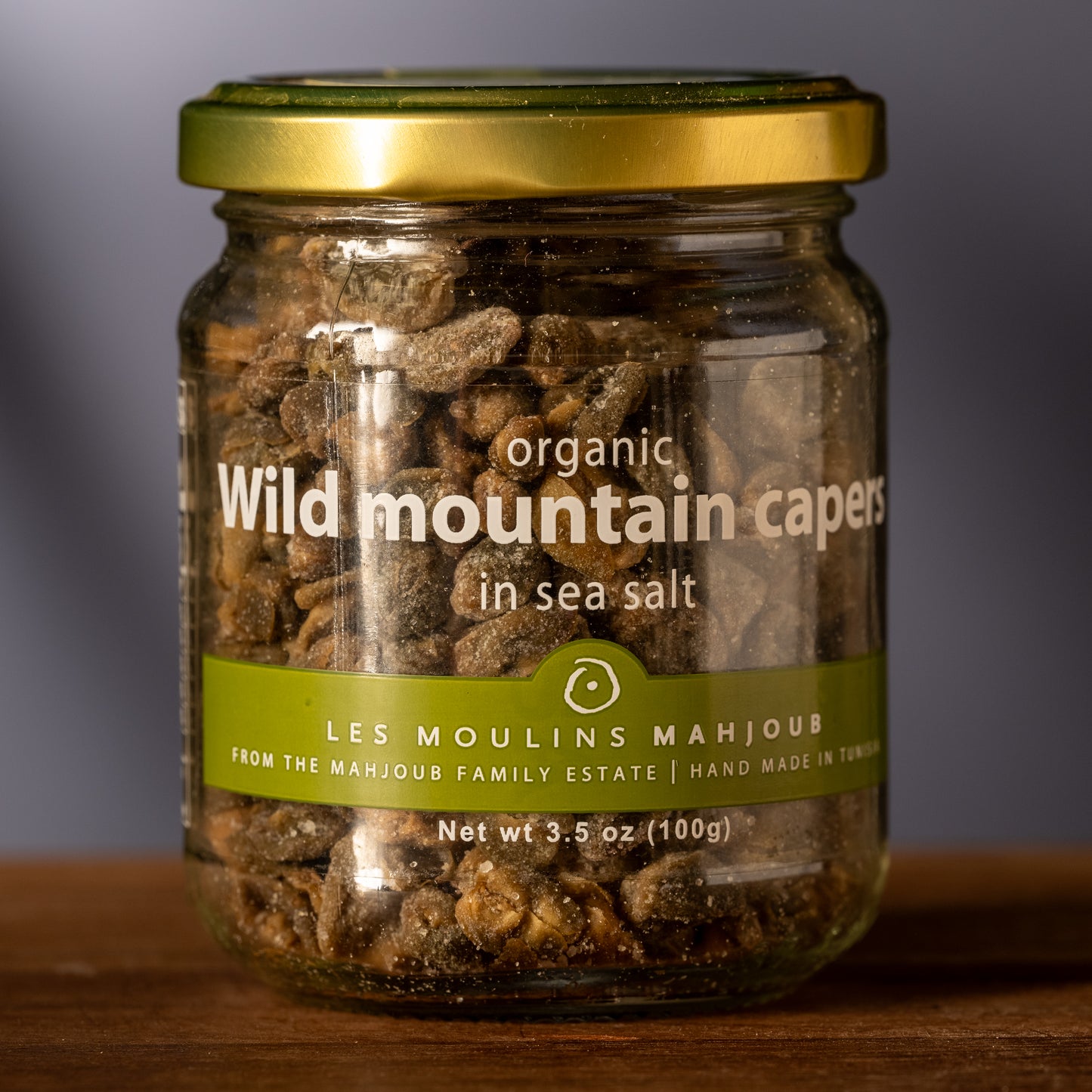 Jar of organic wild mountain capers with a gold lid on a wooden surface.
