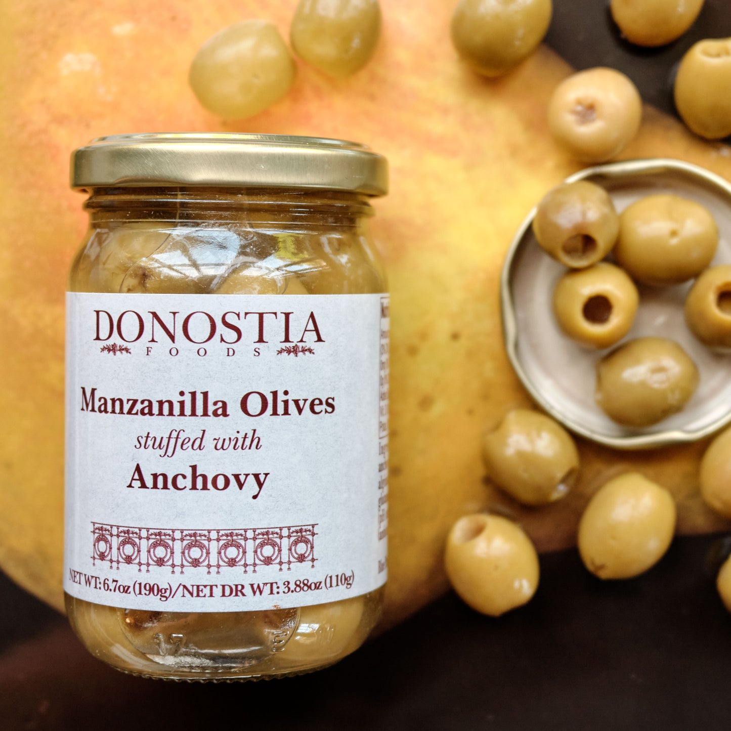 Jar of Donostia Manzanilla Olives stuffed with Anchovy surrounded by olives on  surface.