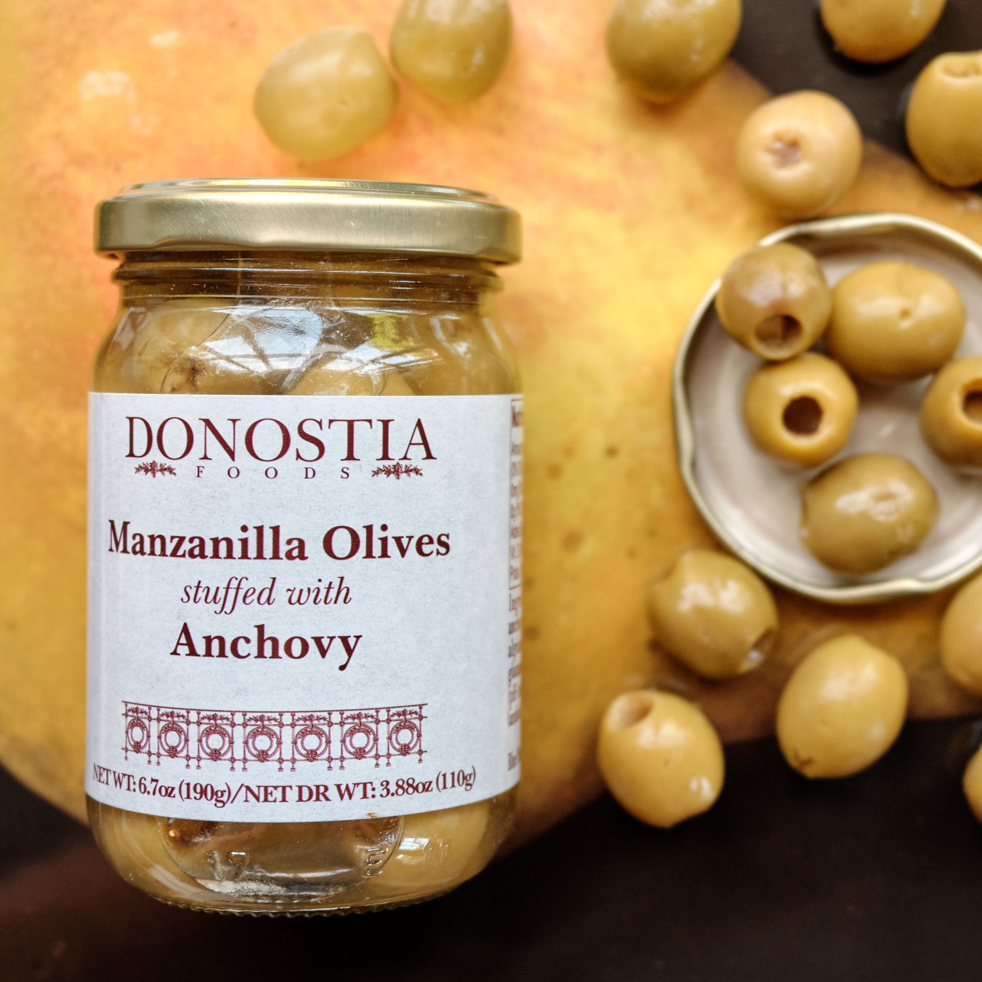 Jar of Donostia Manzanilla Olives stuffed with Anchovy surrounded by olives on  surface.