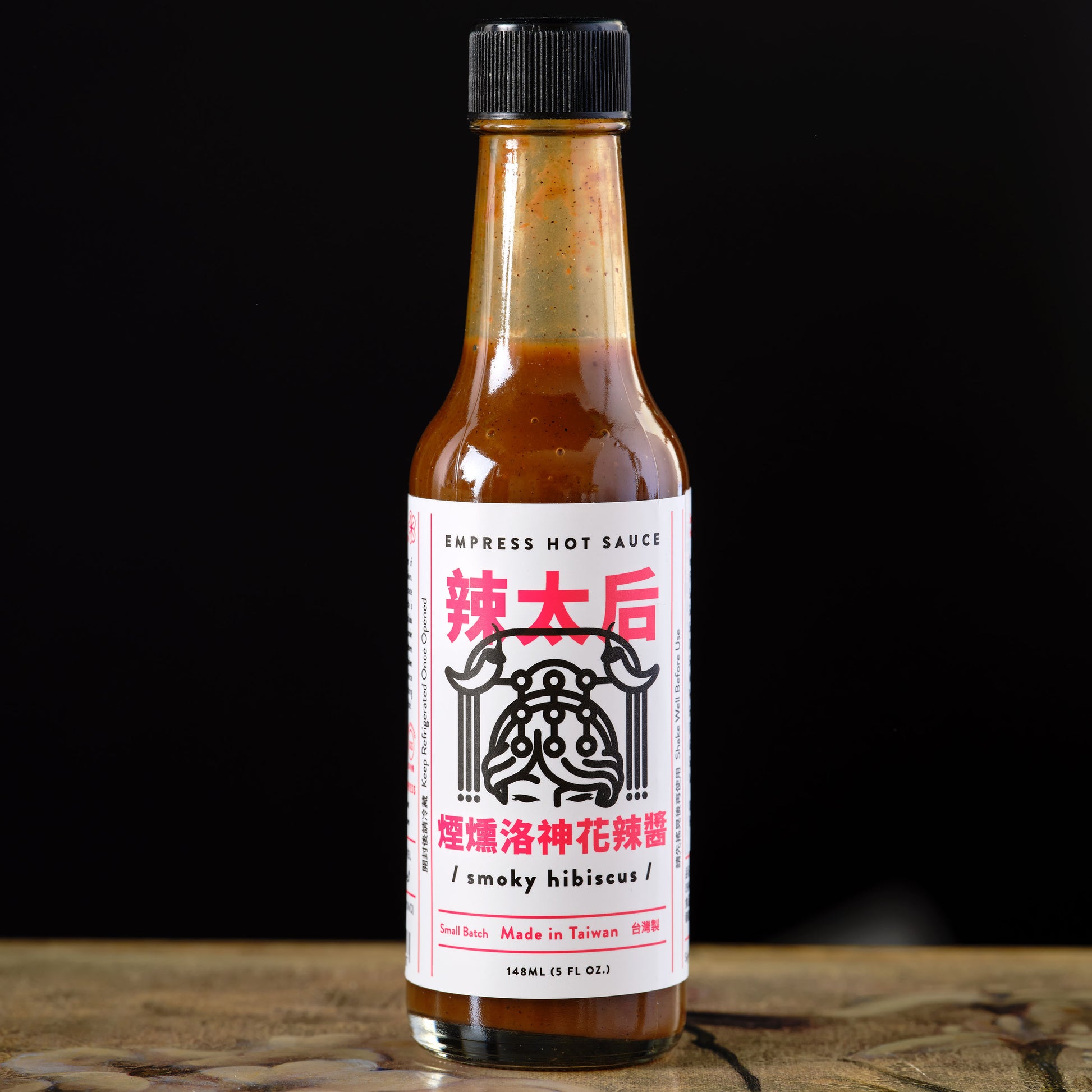 Hot sauce bottle with a label on a wooden surface and black background Empress Smoky Hibiscus Hot Sauce