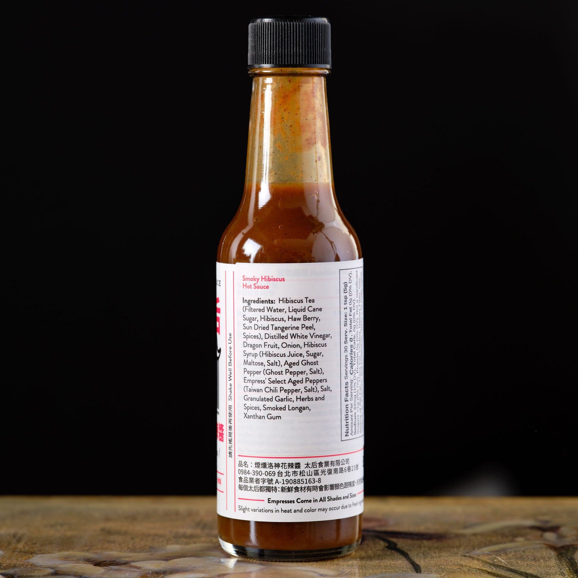Empress Smoky Hibiscus Hot Sauce Bottle of hot sauce with a label on a wooden surface and black background