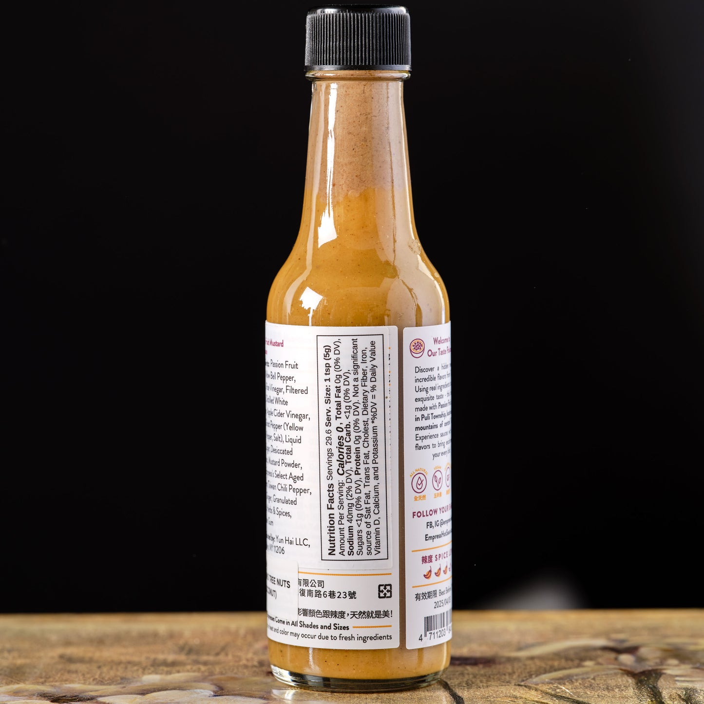 Bottle of hot sauce with a black cap on a wooden surface and dark background Empress Passion Fruit Mustard Hot Sauce