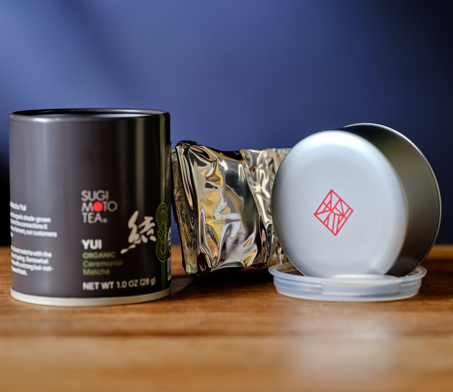 Tea packaging including a black canister, gold leaf, and white container with red logo on a wooden surface.